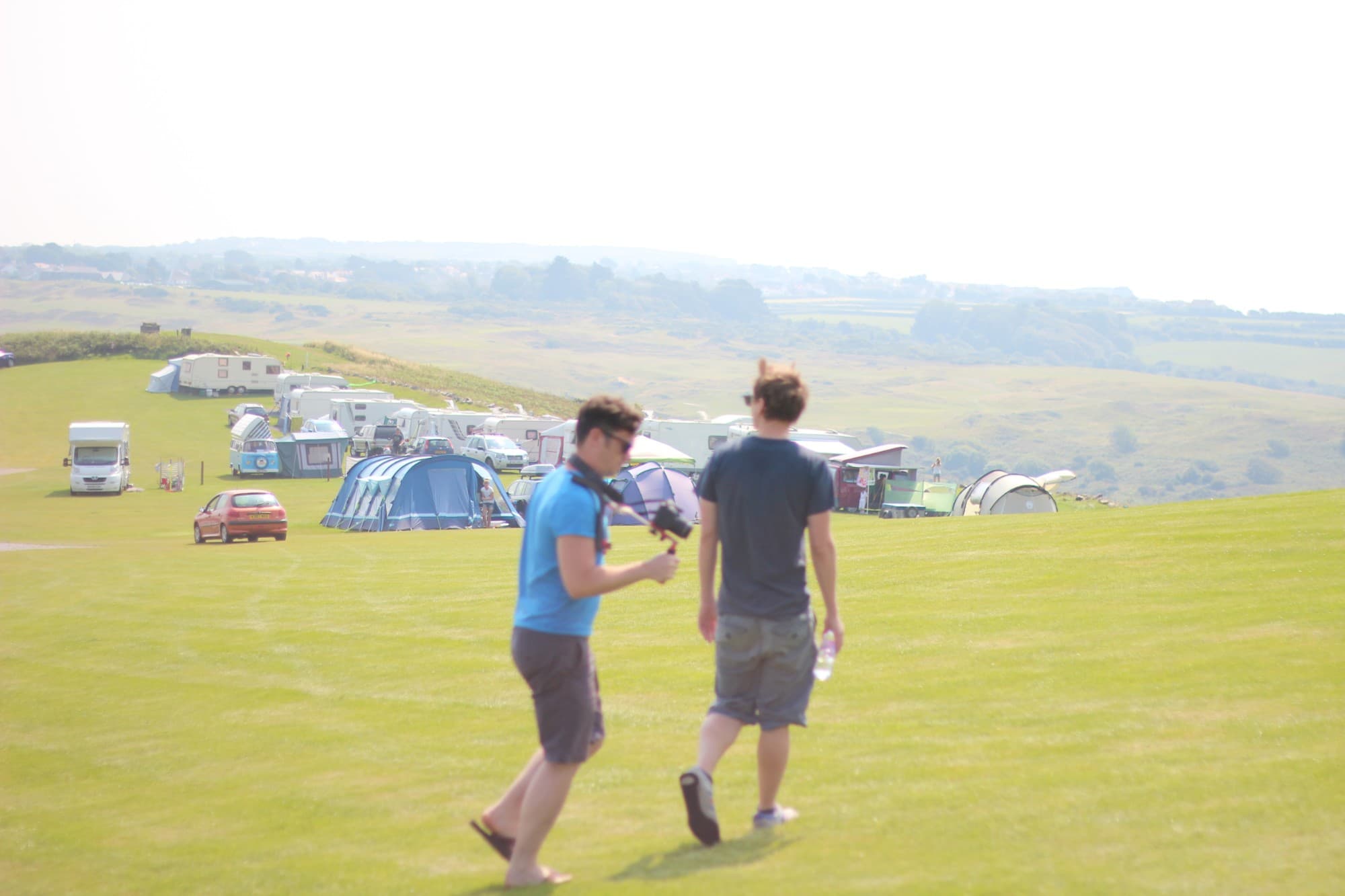 James and Eifion walking across a clifftop campsite field with filming kit on the 2014 road trip