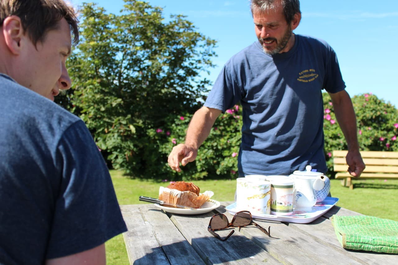 A campsite owner serving tea and homemade cake at a picnic table